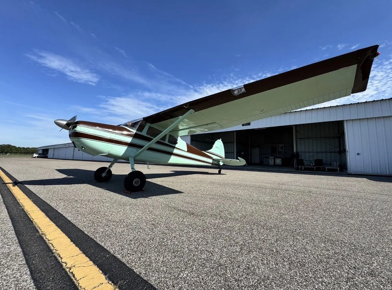 A Cessna 170B aircraft with brown and light green paint scheme parked on an asphalt apron in front of a hangar.