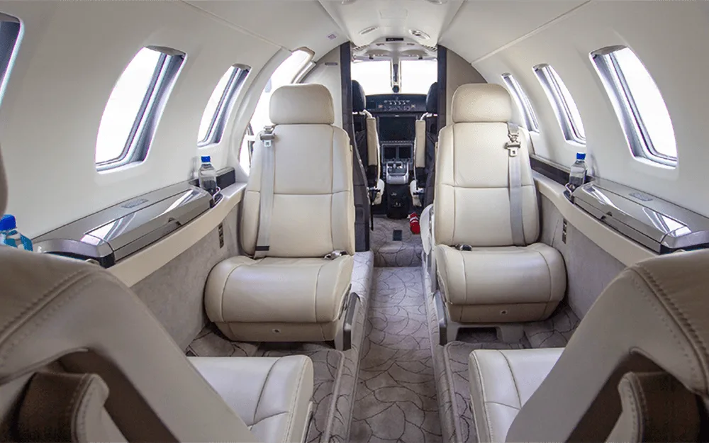 The aircraft cabin interior features light-colored leather seats, a patterned carpet, and a view towards the cockpit.