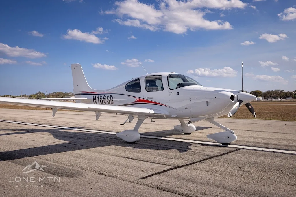 A front-side view of the white N186SB single-engine aircraft with red and black stripes, parked on an airfield.