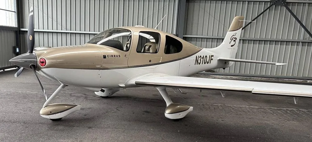 A Cirrus SR22 G3 aircraft with a two-tone tan and white paint scheme is parked inside a hangar, viewed from the front-left.