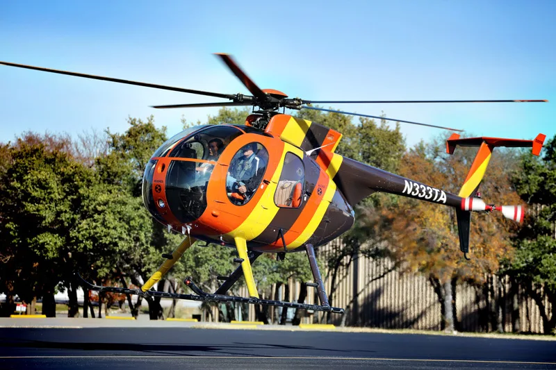 A Hughes 369HS helicopter with orange, yellow, and black stripes takes off from an airfield.