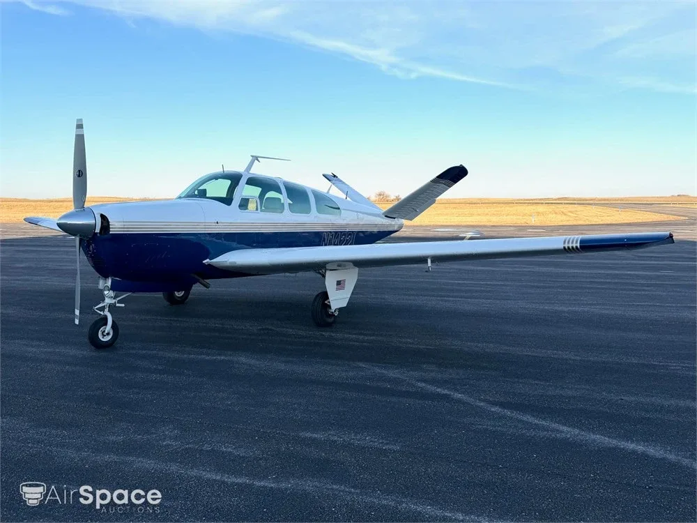 Left side view of a blue and white Beechcraft Bonanza parked on an asphalt runway under a clear sky.