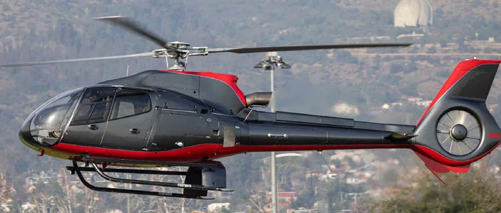 A dark grey and red helicopter flies over a hilly landscape with buildings in the background.