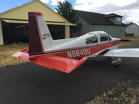 A rear-side view of the white and red Grumman American AA-5 Traveler (N9840U) parked near a yellow hangar.