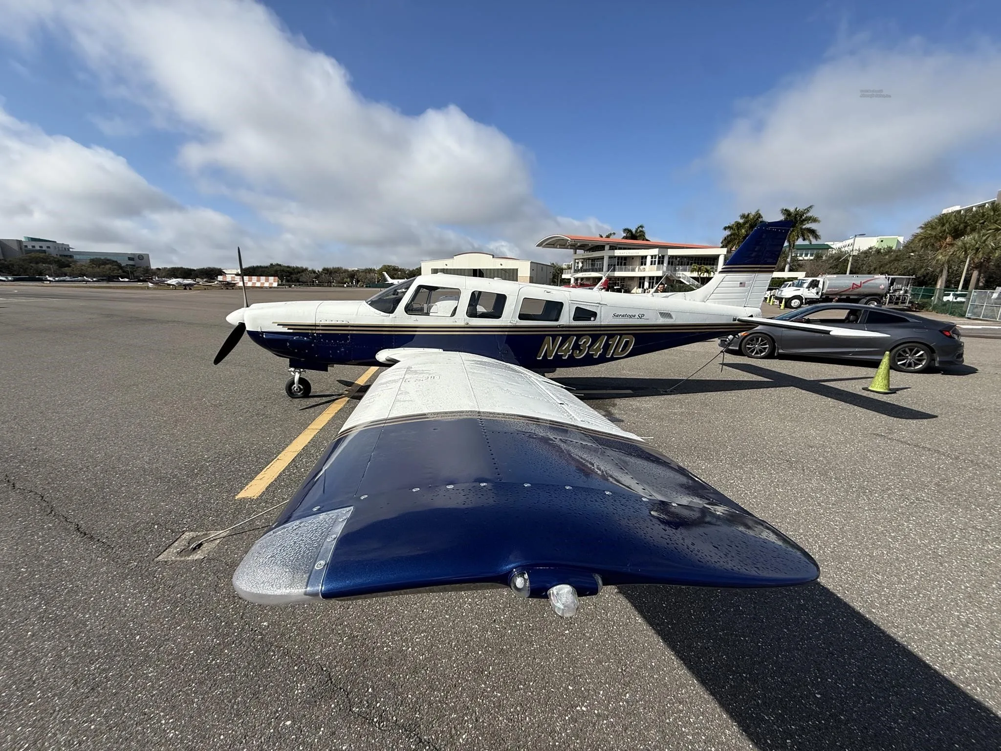 A Piper Saratoga SP (N4341D) in blue and white paint scheme parked on an airfield next to a grey car.