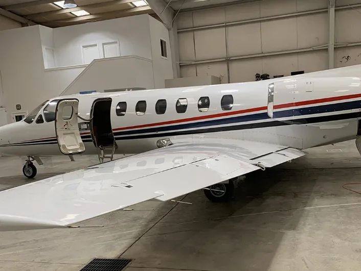 A close-up side view of a white private jet with red and blue stripes, showing the open main door and wing, inside a hangar.