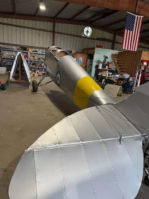 The silver tail section of a biplane with a yellow stripe and roundel is visible in a hangar.