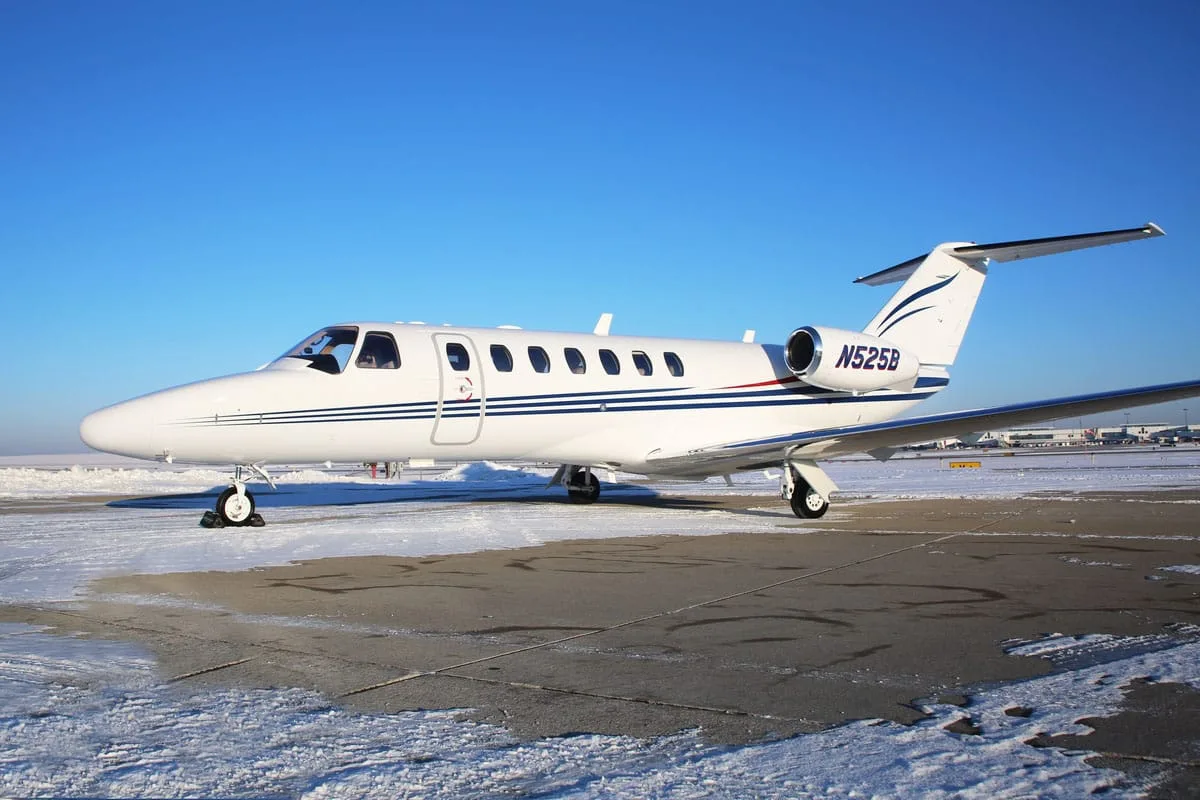 A white Cessna CitationJet with blue stripes, registration N525B, parked on a snowy tarmac under a clear blue sky.
