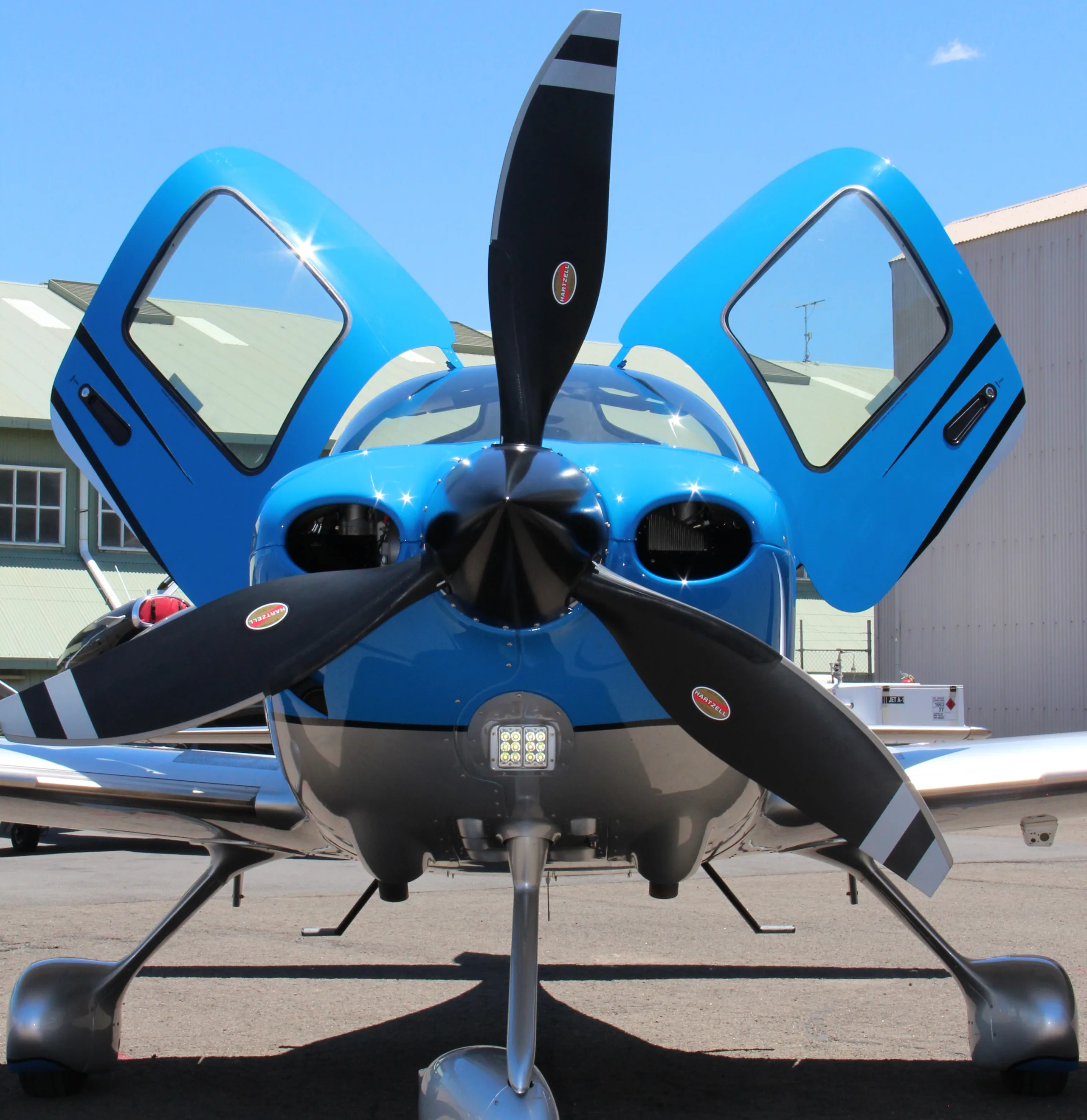 A blue and silver single-engine propeller aircraft with open gull-wing doors is parked on an tarmac.