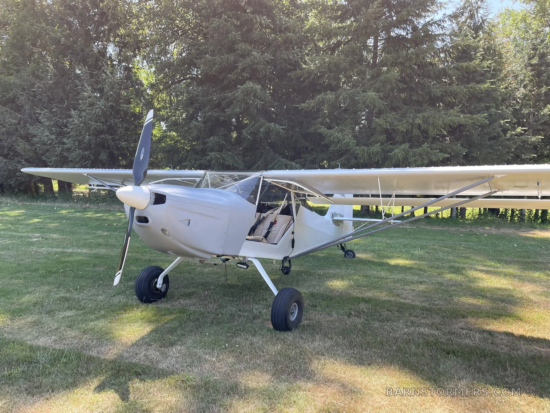 A light grey single-engine aircraft with a high wing and large tires is parked on a grassy field with tall green trees in the background.