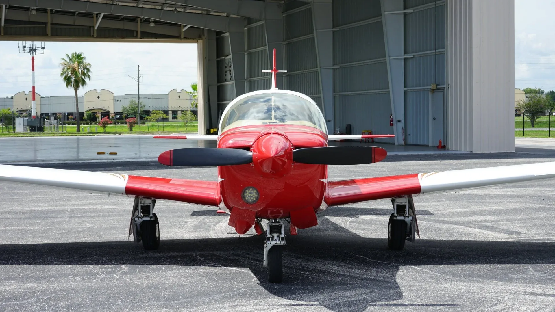 A head-on view of a red and white single-engine aircraft with a black propeller, parked in front of a hangar.