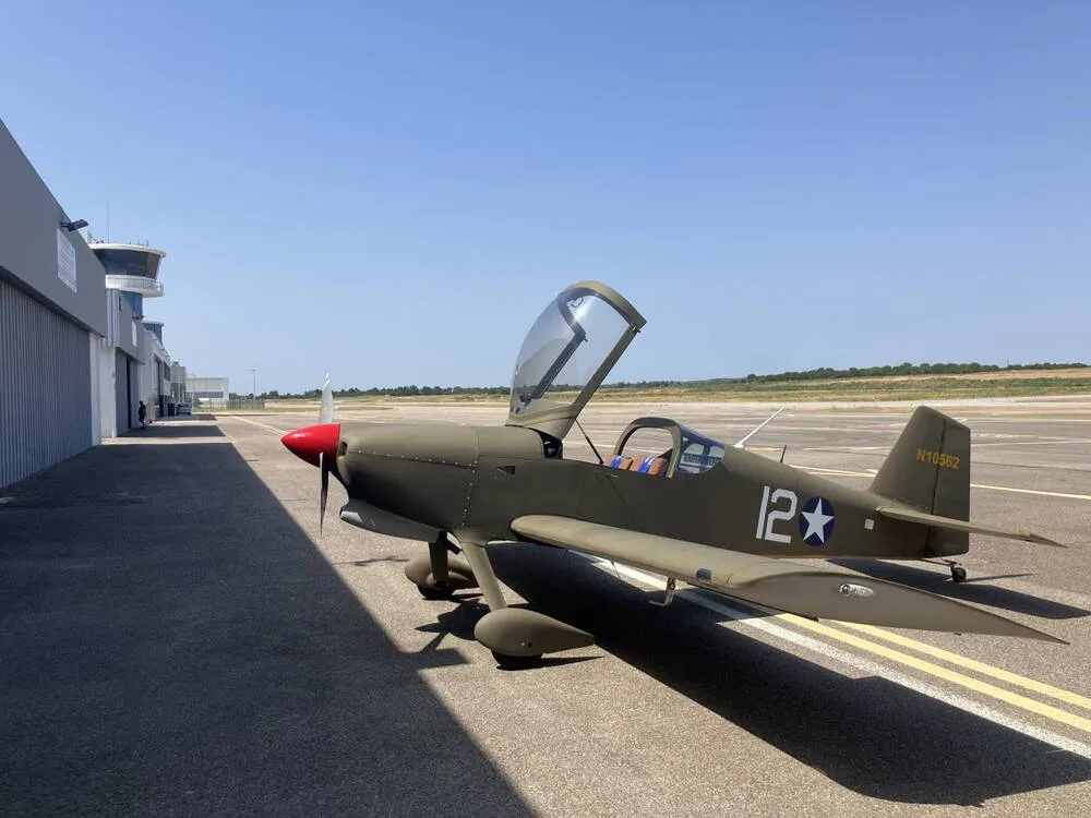 A green single-engine aircraft with a red nose and "N10562" on the tail, parked on an airfield with a control tower in the background.