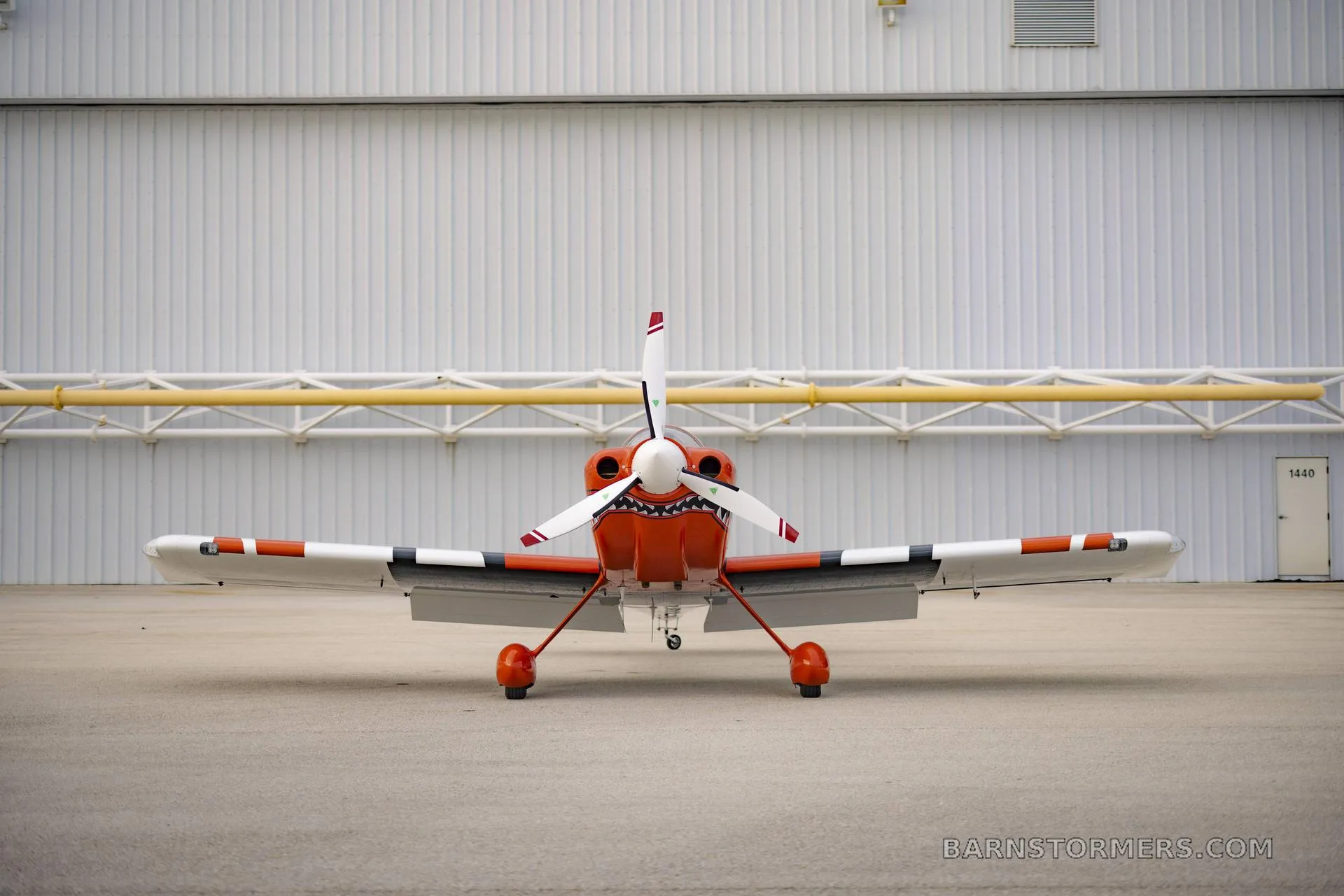 A head-on view of an orange and white single-engine aircraft with a shark mouth paint scheme, parked in front of a hangar.