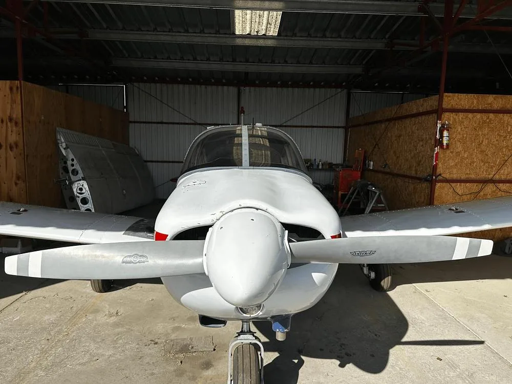 A head-on view of a white Piper Cherokee C aircraft with a two-blade propeller, parked in a hangar.