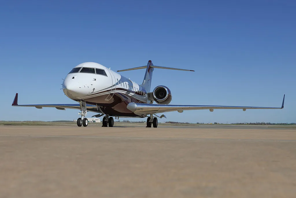 A Bombardier Global Express business jet with a white and maroon paint scheme, registration N919EM, parked on a tarmac under a clear blue sky.