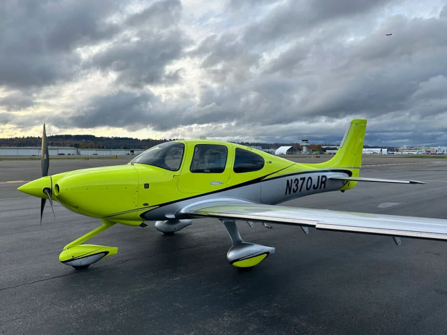 A bright yellow and silver Cirrus SR22 aircraft with registration N370JR parked on an airport tarmac under cloudy skies.