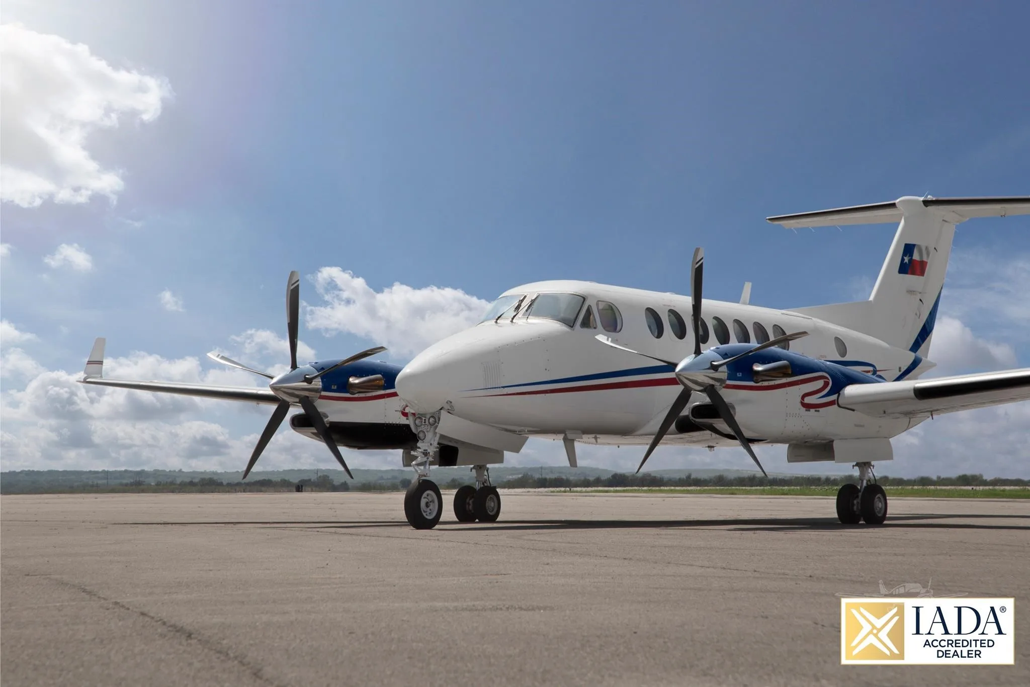 A white Beechcraft King Air twin-turboprop with blue and red stripes and a Texas flag on its tail sits on an airfield under a partly cloudy sky.