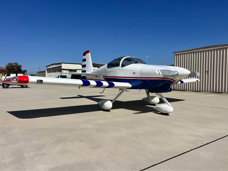A white, blue, and red single-engine aircraft with a bubble canopy and striped wings, parked on an airfield with hangars in the background.