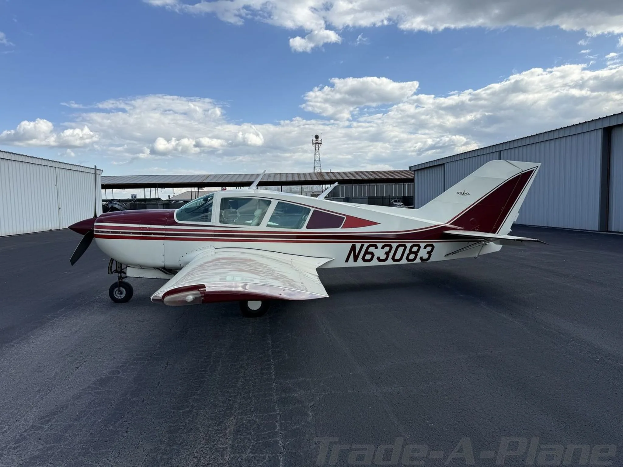 Side view of a white and maroon Bellanca Viking N63083 parked on a tarmac with hangars in the background.
