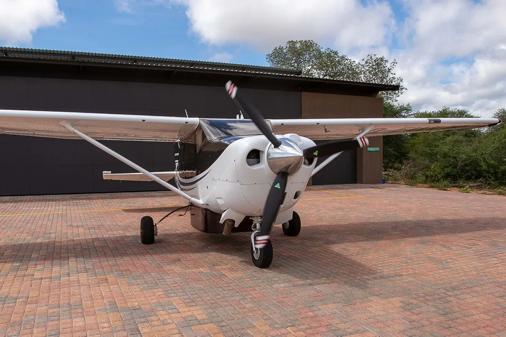A white and black single-engine propeller aircraft is parked on a brick-paved area in front of a dark hangar under a blue sky.
