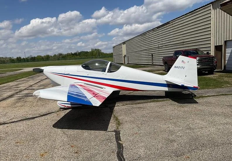 A side view of a white single-engine aircraft with red and blue stripes, parked on a tarmac next to a hangar and a red pickup truck.