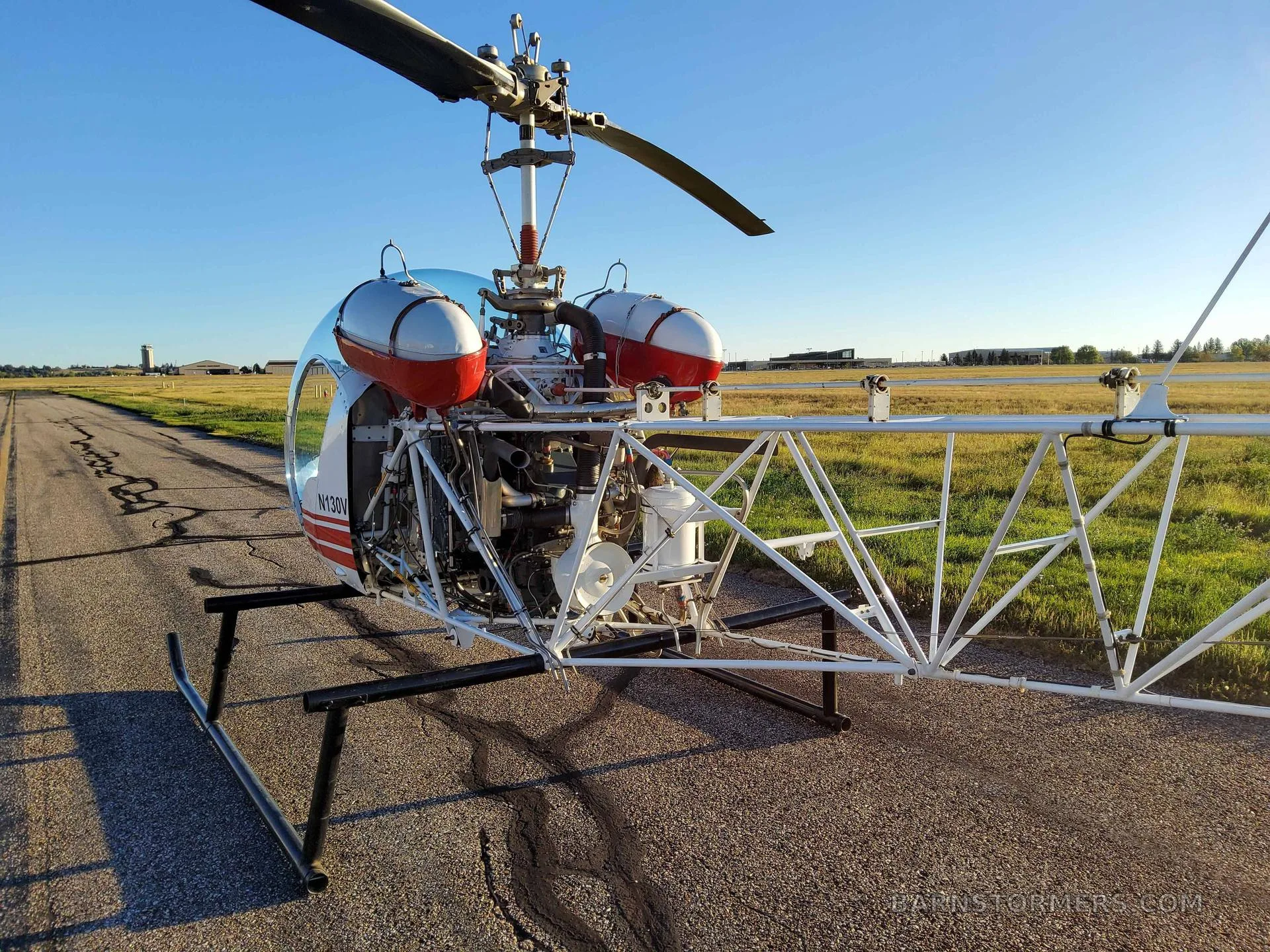 A side view of a white helicopter with an exposed engine and open-truss tail boom, parked on an airfield.