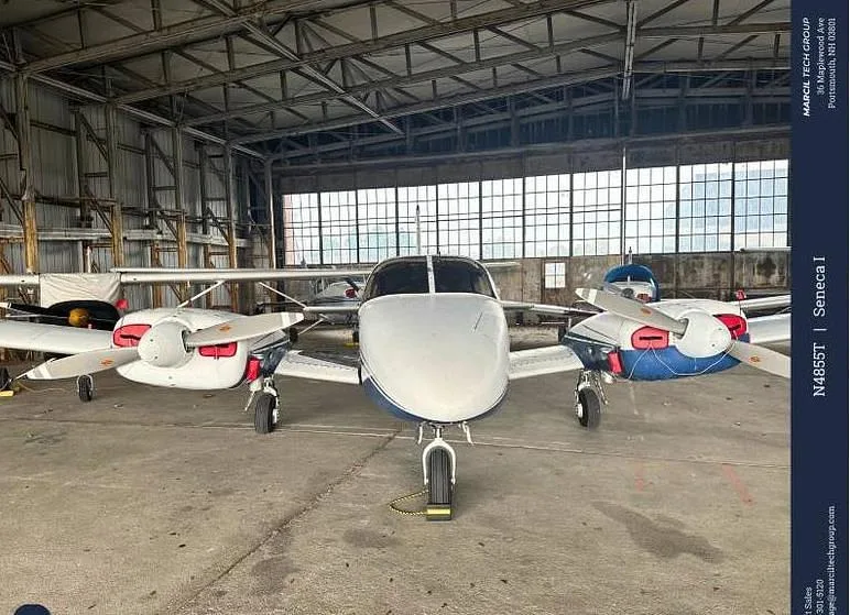 A white and blue Piper Seneca I aircraft is parked head-on inside a hangar, showing its twin engines.