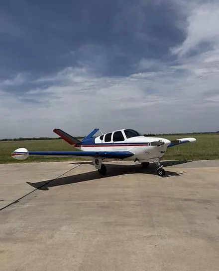 A white single-engine aircraft with blue and red stripes parked on an asphalt runway under a partly cloudy sky.