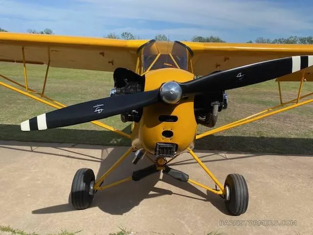 A front view of a yellow Piper Cub-style aircraft with a black propeller, parked on a concrete pad.