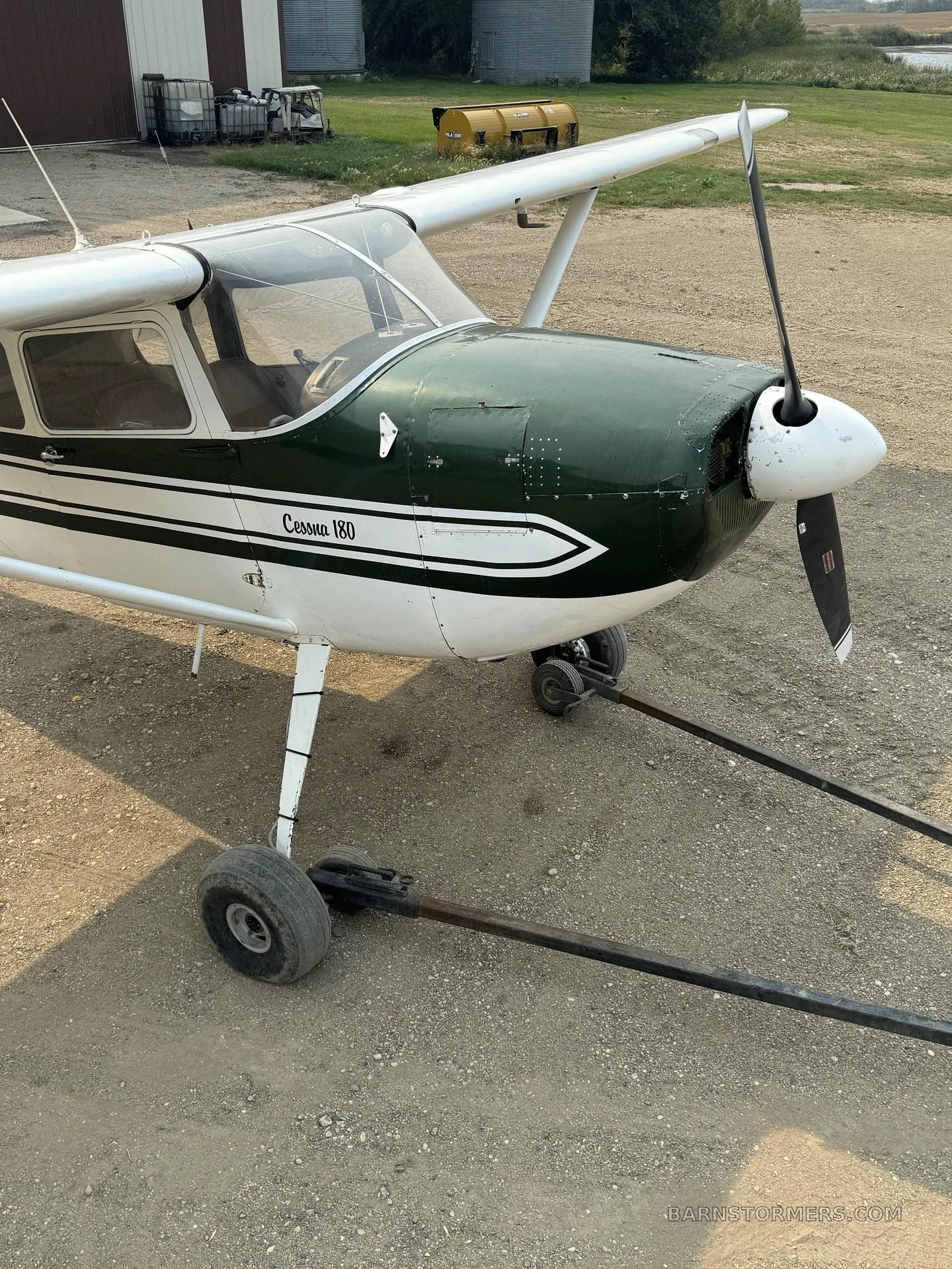 A front-right view of a white and green Cessna 180 aircraft parked on a gravel lot.