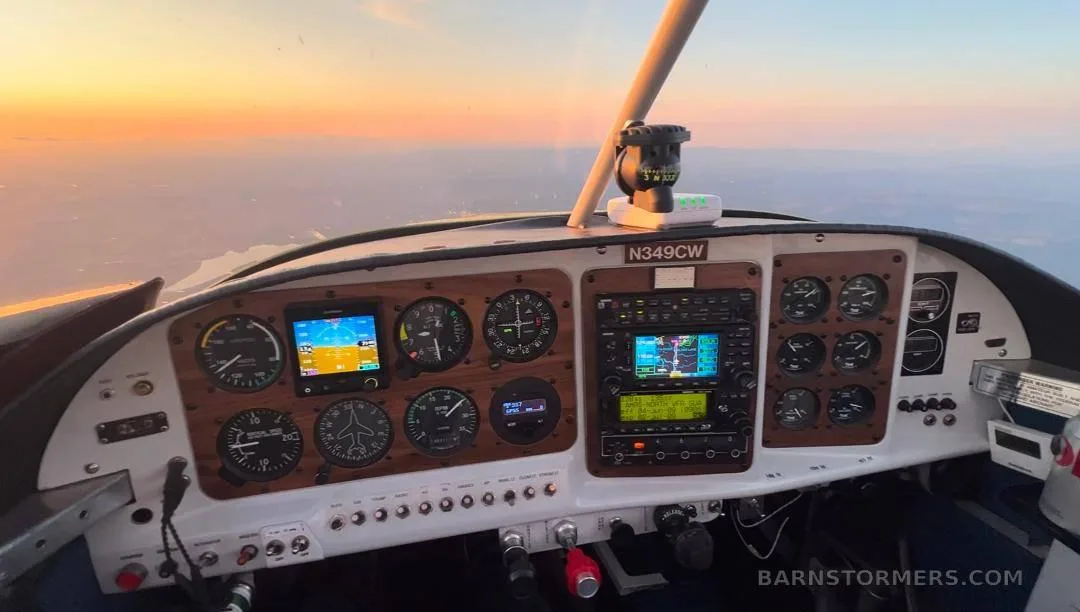 The cockpit instrument panel of an aircraft, featuring analog gauges and digital displays, with a sunset visible through the windshield.