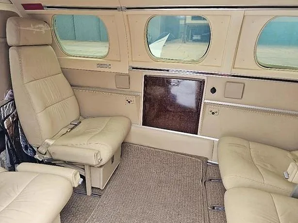 The aircraft cabin interior with tan leather seats, a carpeted floor, and a dark wood-paneled cabinet.