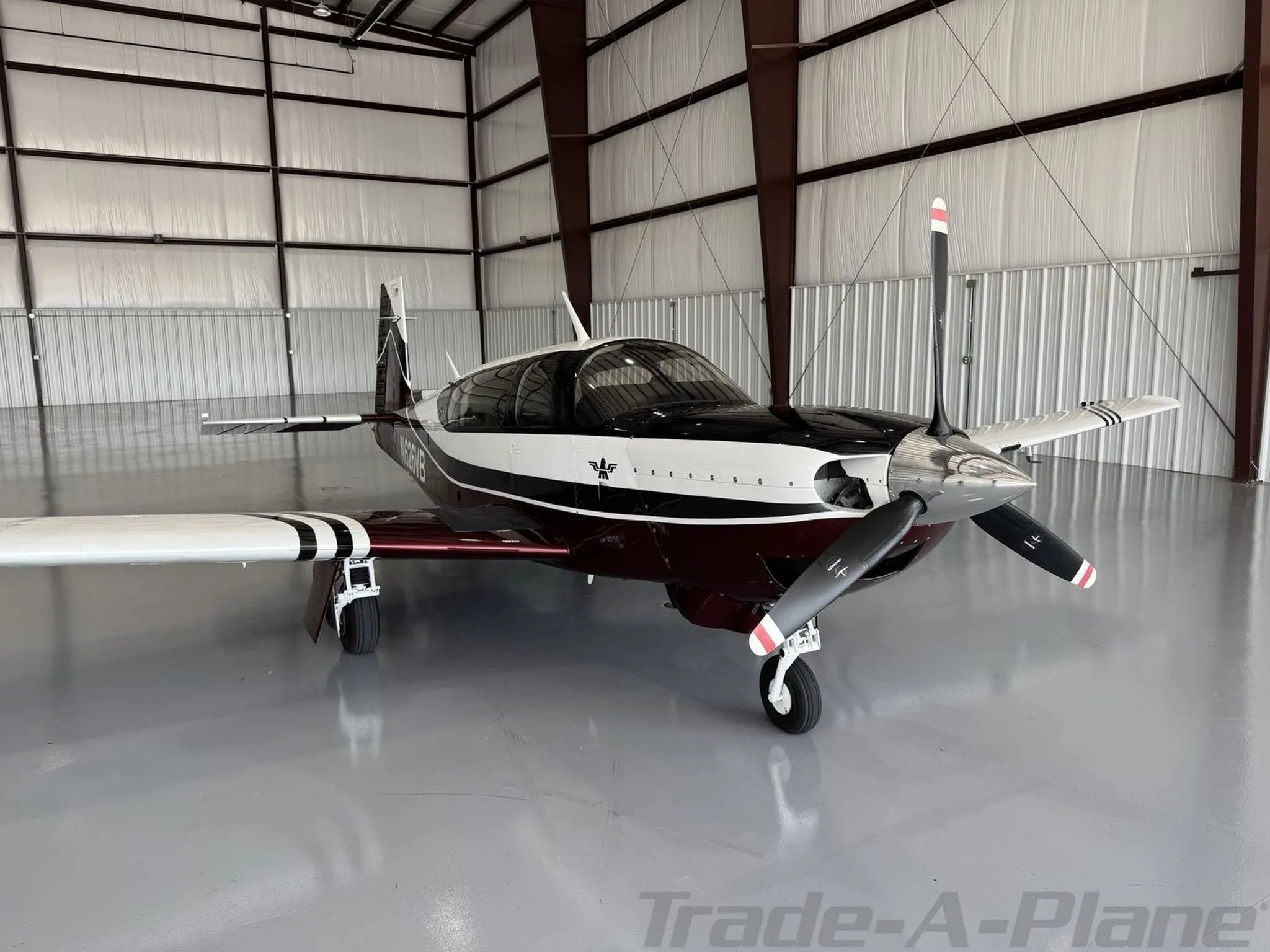 A Mooney aircraft with a maroon, white, and black paint scheme is parked in a hangar, viewed from the front right.
