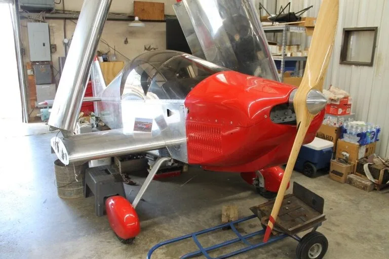 A silver and red experimental aircraft with a wooden propeller is shown in a workshop, with its canopy open and wings partially folded.