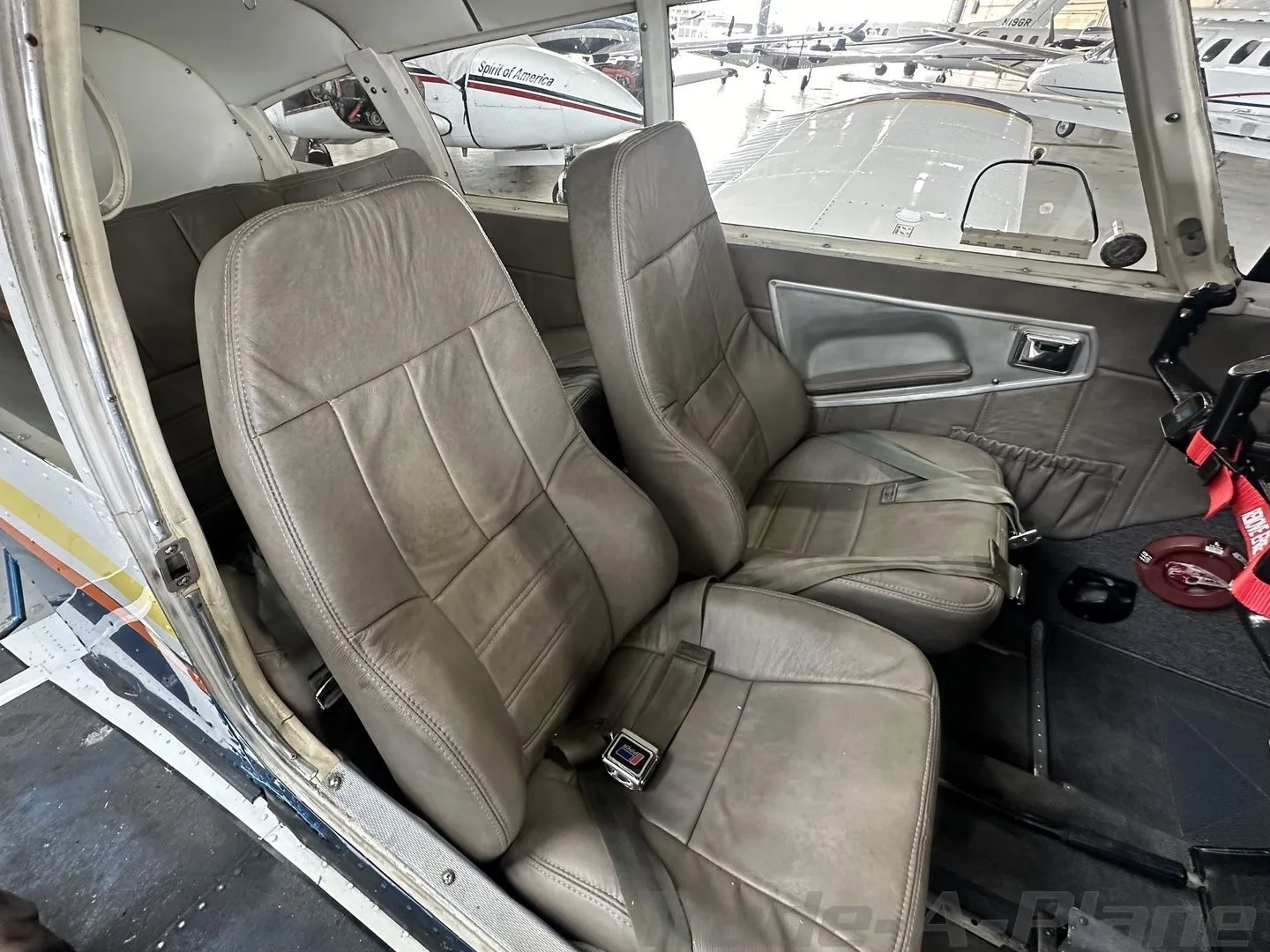 The tan leather front seats of a light aircraft, N3905T, with seatbelts, viewed from inside a hangar.