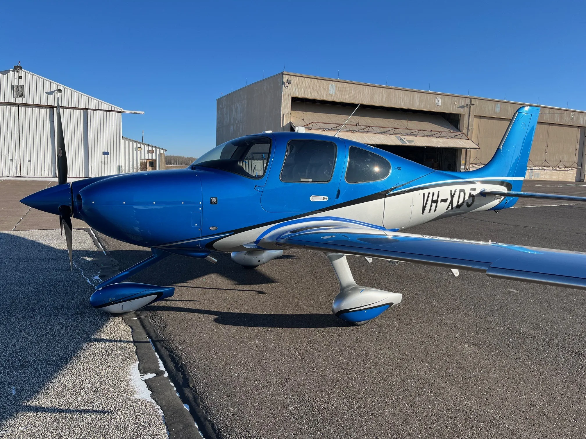 A blue and white single-engine Cirrus SR22 aircraft with registration VH-XD5 parked on an airfield next to hangars.