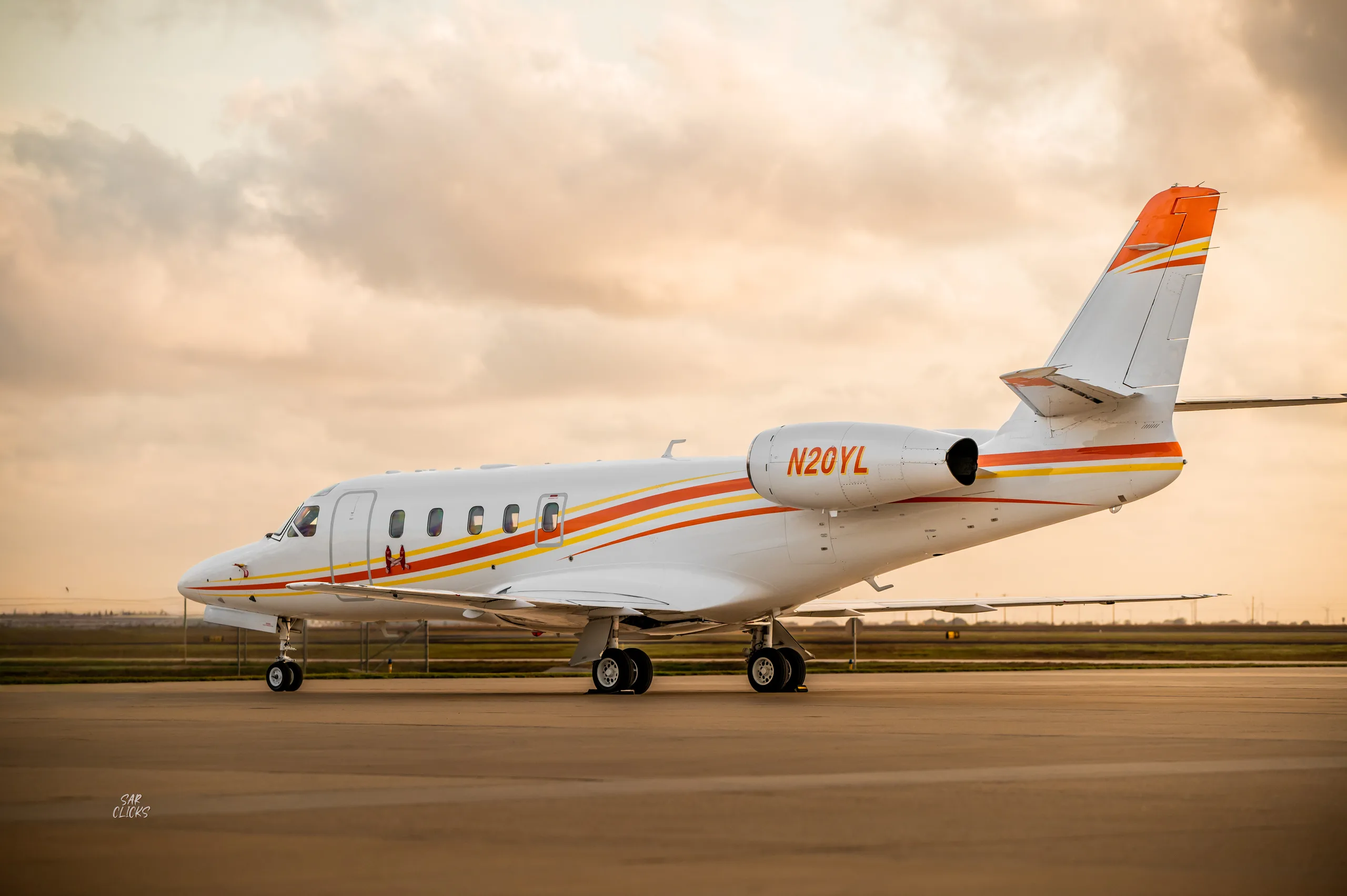 A white business jet with orange and yellow stripes, tail number N20YL, sits on an airfield at sunset.