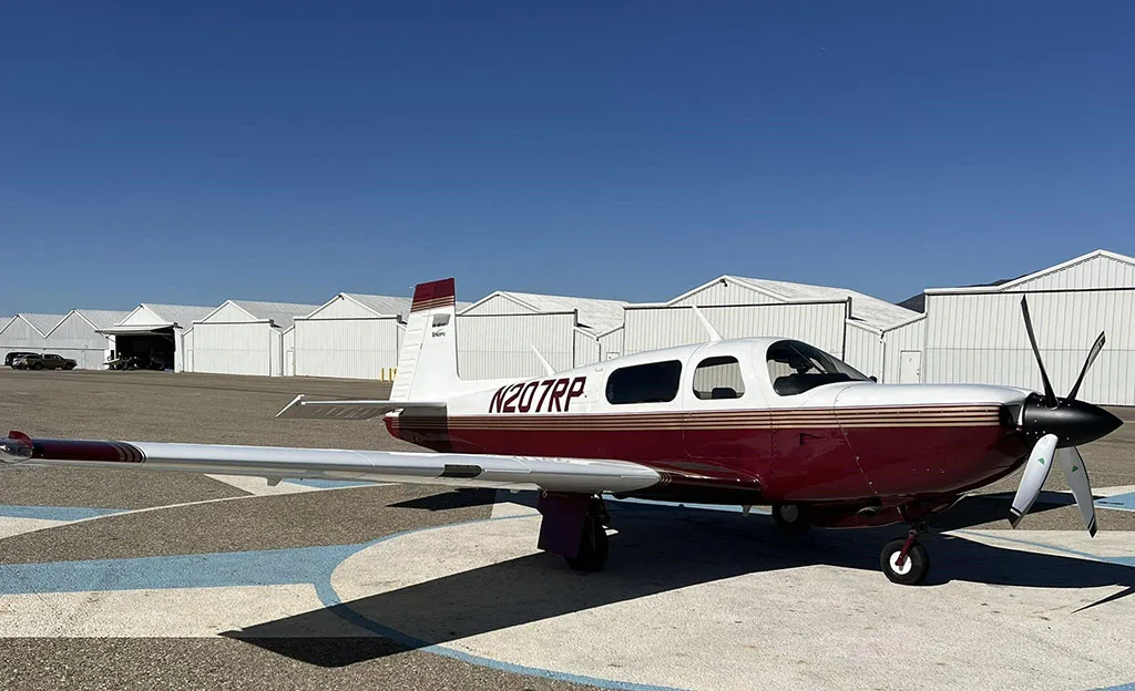 A white and maroon single-engine propeller aircraft, N207RP, is parked on an airfield with hangars in the background, viewed from the right side.