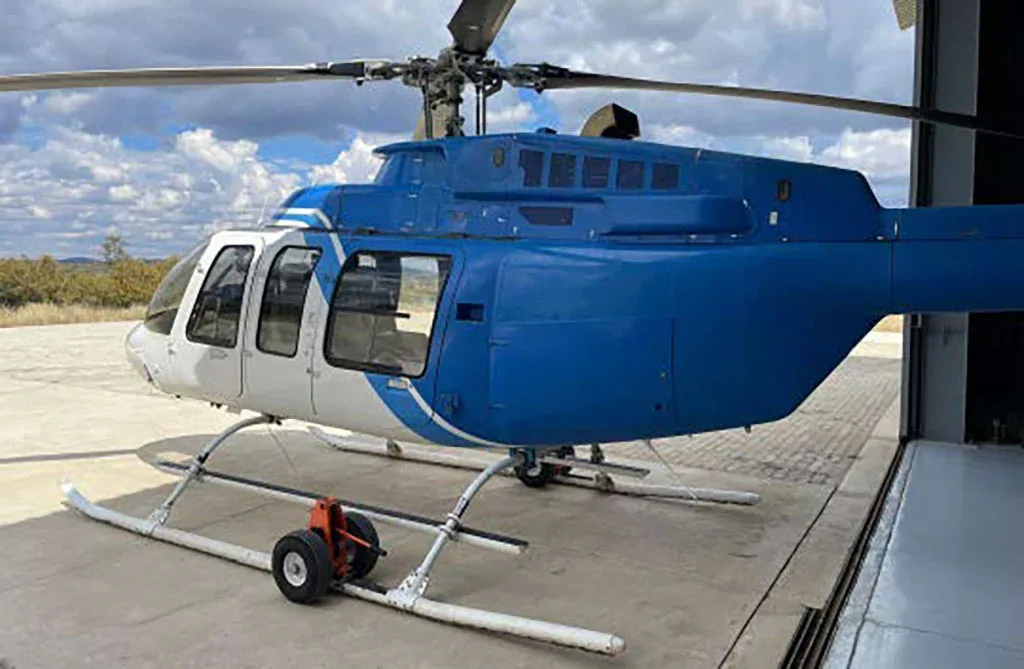 A blue and white helicopter, viewed from the side, parked on concrete with a hangar in the background.