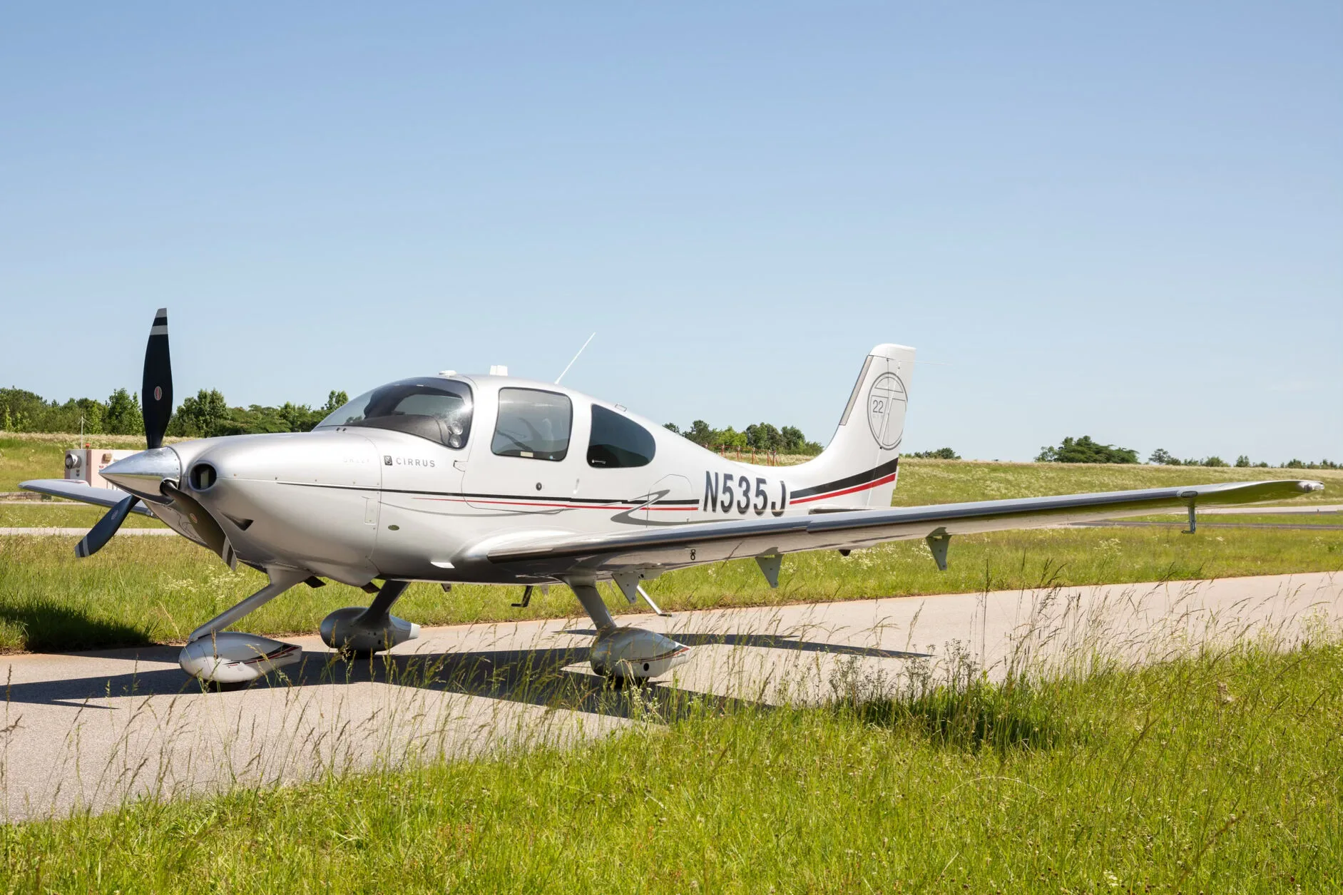 A silver Cirrus SR22T aircraft with registration N535J is parked on an asphalt strip next to green grass under a clear blue sky.