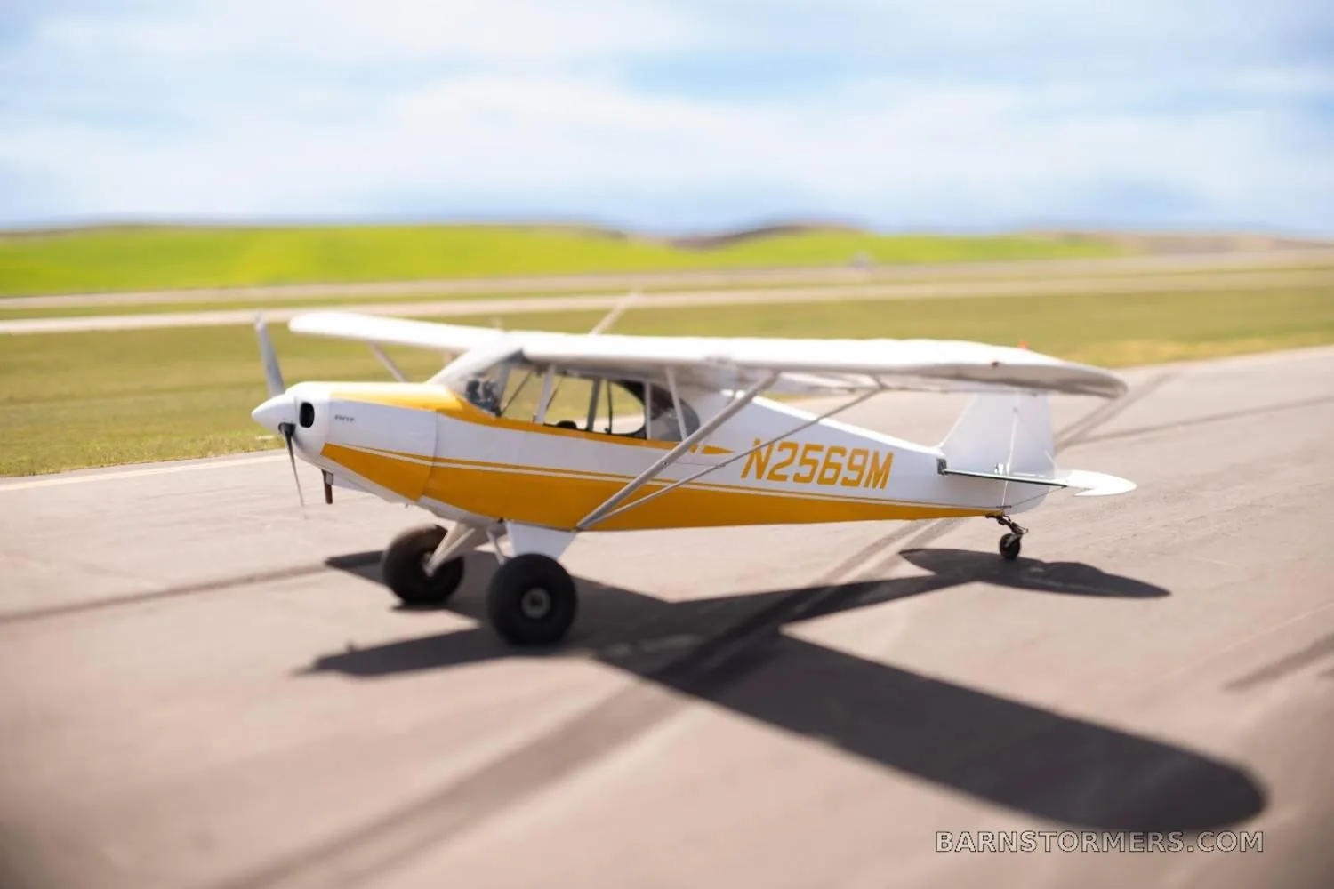 A yellow and white single-engine taildragger aircraft (N2569M) with large tires on an asphalt runway, with green hills in the background.