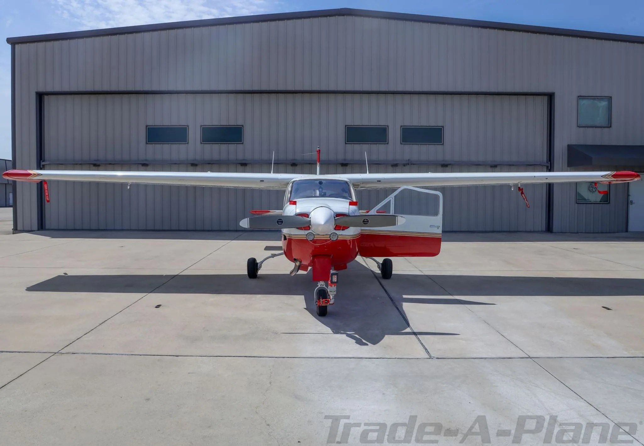 A red and white Cessna 177RG Cardinal with its door open, parked in front of a hangar.