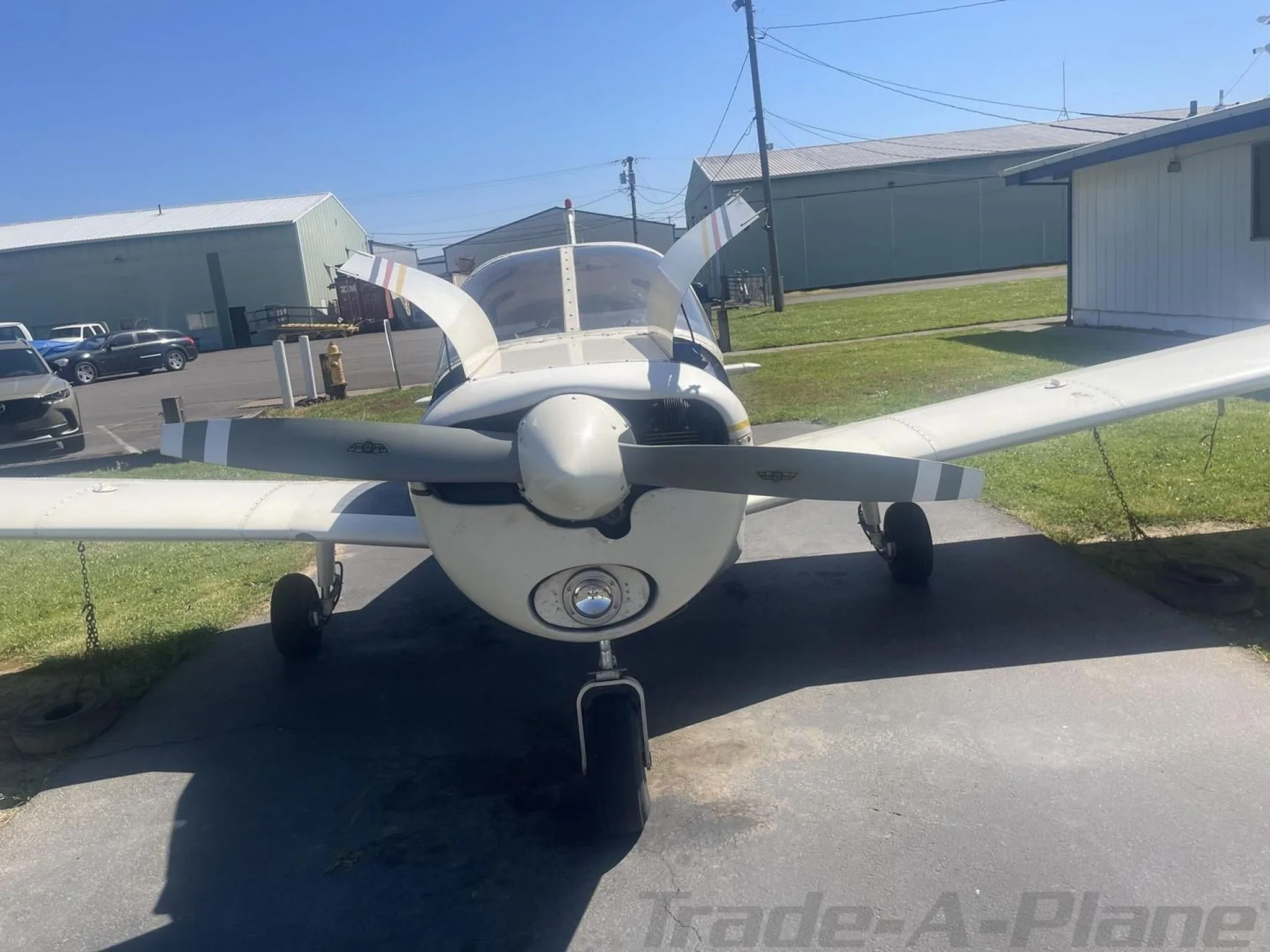 Front view of a white single-engine propeller aircraft with a grey propeller, parked on asphalt in front of hangars.