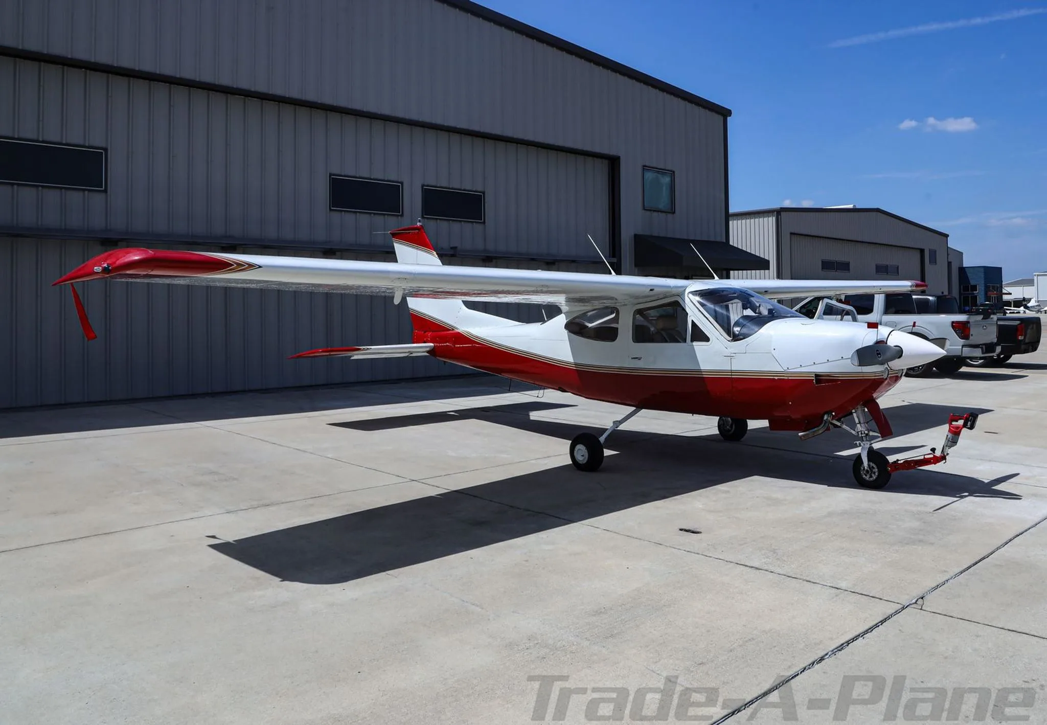 A red and white Cessna 177RG Cardinal with gold stripes parked on an airfield next to a hangar.