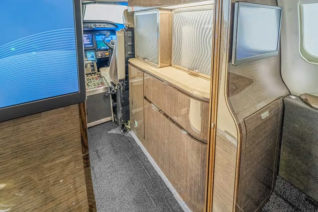 A view of the galley area in the business jet's cabin, featuring wood-paneled cabinets and a screen, with the cockpit visible in the background.