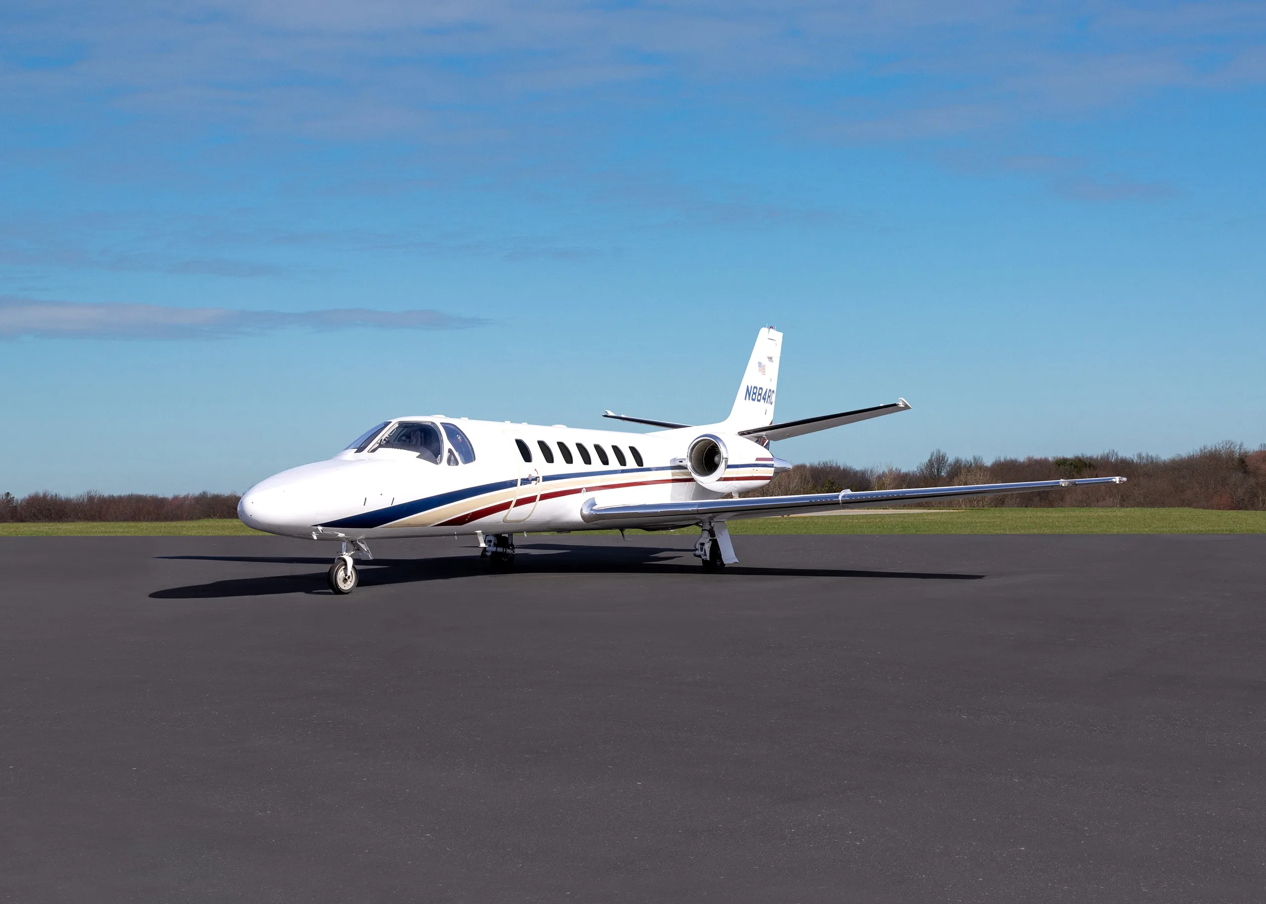 A white business jet with blue and red stripes, registration N884RC, parked on an asphalt tarmac under a blue sky.