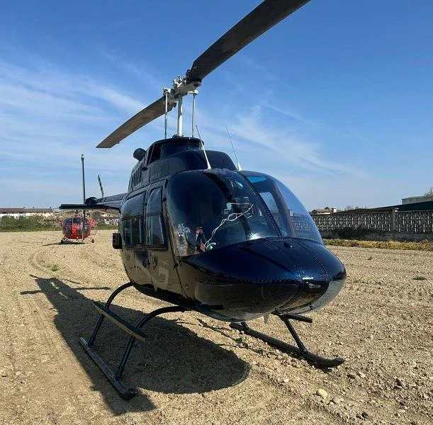 A dark blue Bell 206 helicopter is parked on a dirt field, viewed from the front-right, with another red helicopter in the distance.