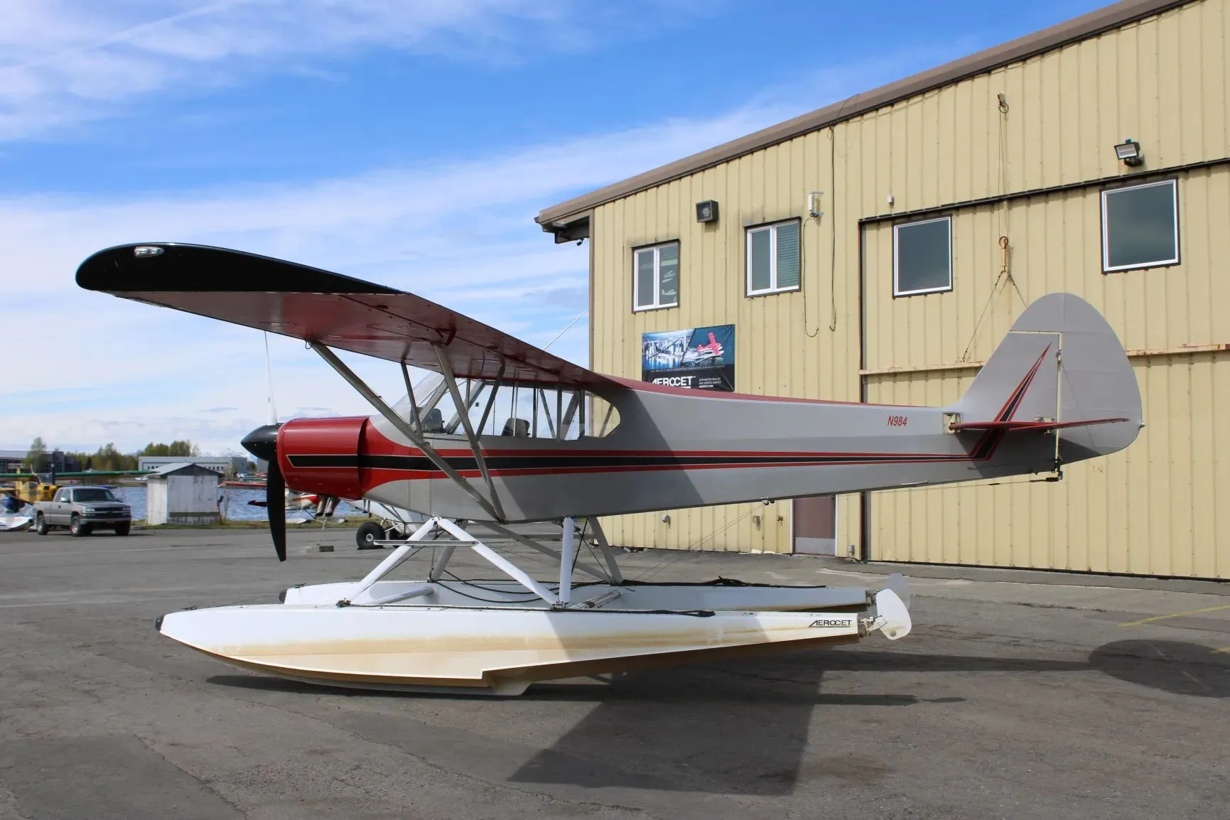 A side view of a red and grey floatplane with N984 on the tail, parked next to a yellow hangar under a blue sky.