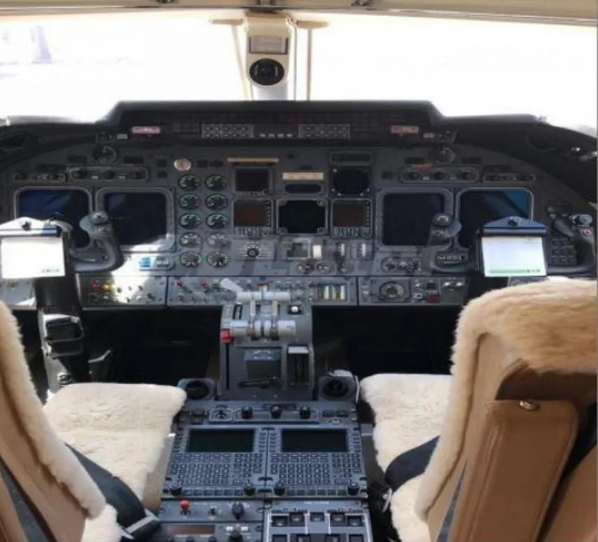 The cockpit of a private jet, showing the instrument panel, controls, and two pilot seats with sheepskin covers.