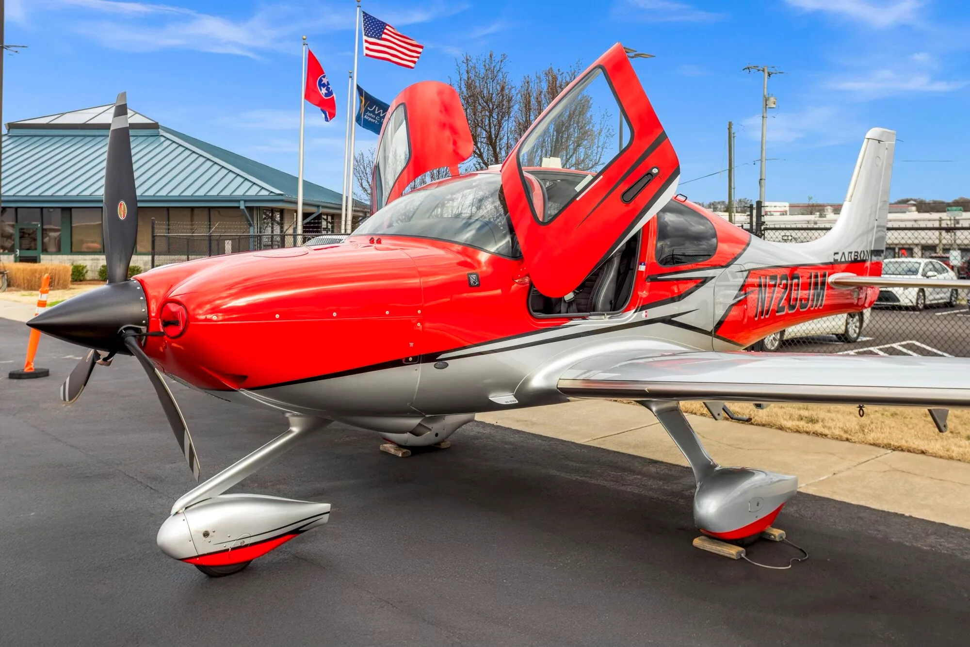 A front-quarter view of a red and silver Cirrus SR22 aircraft with its gull-wing doors open, parked on an airport tarmac.